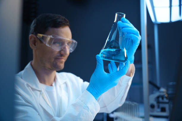 A young white chemist looks at a laboratory flask with blue liquid in it, sitting in a secret laboratory, we see him from the distance