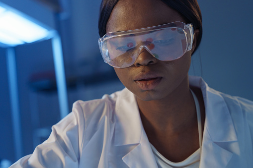A close-up of a young concentrated African-American scientist in protective eyewear, sitting at her desk in a modern laboratory