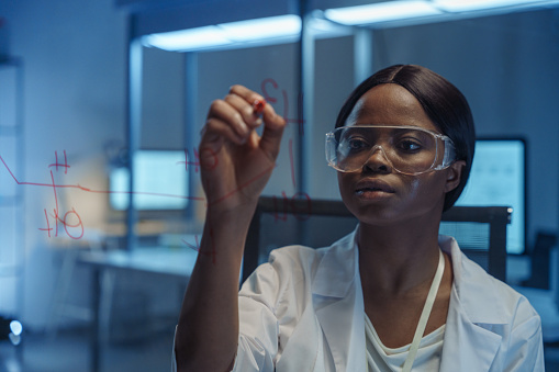 A young female African-American chemist behind a glass board, writing chemical formulas on it