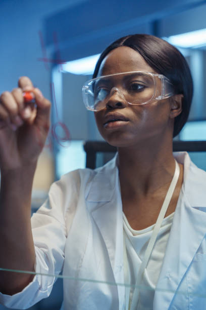 유리 판에 공식을 쓰는 젊은 사려 깊은 아름다운 아프리카 계 미국인 과학자, 우리는 그 보드 뒤에서 그녀를 봅니다. - genetic research chemist writing science 뉴스 사진 이미지