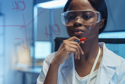 A young smart African-American scientist thinking on solution of a chemical reaction written on  a glass board in front of her in a modern laboratory
