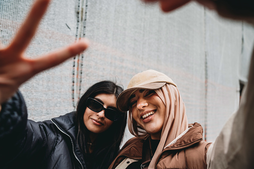 Two young adult girls are making funny gestures to the camera. One of them is wearing a fashionable hijab with an hat.