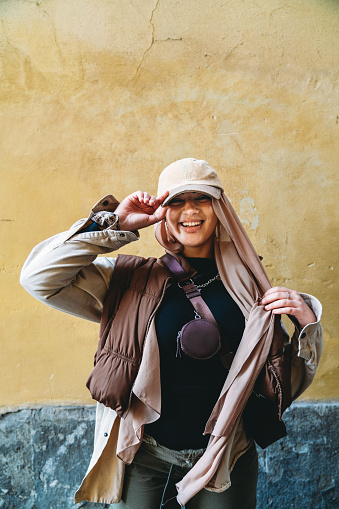 Portrait of a young adult woman against a yellow background. She's wearing a fashionable hijab with an hat.