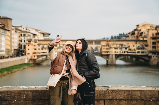 Two friends are taking a selfie on Santa Trinita bridge with Ponte Vecchio in the background