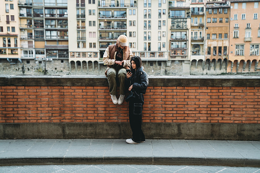 Two friends are hanging out in Florence, Italy. One of them is sitting on a low wall in Lungarno street.