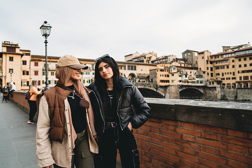 Two friends are walking together in Florence's Lungarno street. One of them is wearing a fashionable hijab with an hat.