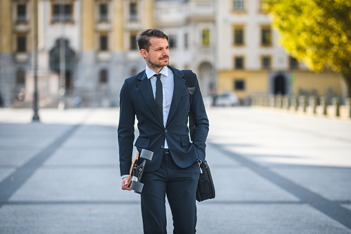 Handsome young businessman, holding a longboard and carrying a messenger bag, hand in pocket, walking down a street of old town, dressed for success. Early morning, slowly going to work. Focus on foreground. Looking away. 3/4 shot.