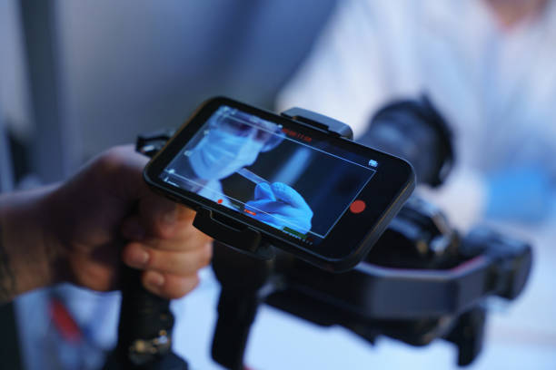 A hand of an operator with a shooting camera and a smart phone screen, showing an image from the camera, where we see a man in a protective mask