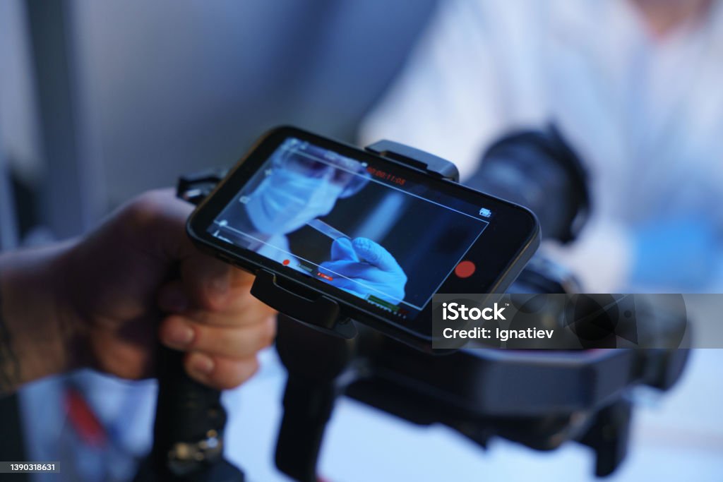 A hand of an operator with a shooting camera and a smart phone screen, showing an image from the camera, where we see a man in a protective mask Movie Stock Photo