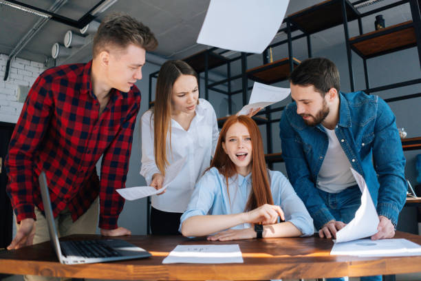 istock Furious sad young business woman under stress throwing documents sitting at desk looking at camera, multi-ethnic colleagues standing near her. 1390319675