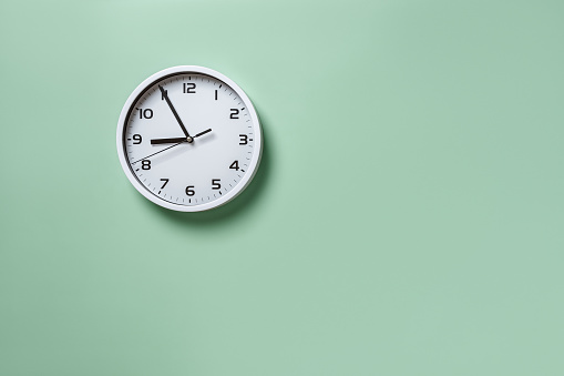 Wall clock hanging on the pale green wall with copy space. Round white clock with black hands. Five minutes to nine. Time measuring, hour and minutes concepts. Time control. Working hours. Front view.