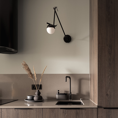Close-up on beige kitchen countertop with decorations, stylish tap and lamp, small sink and wooden furniture