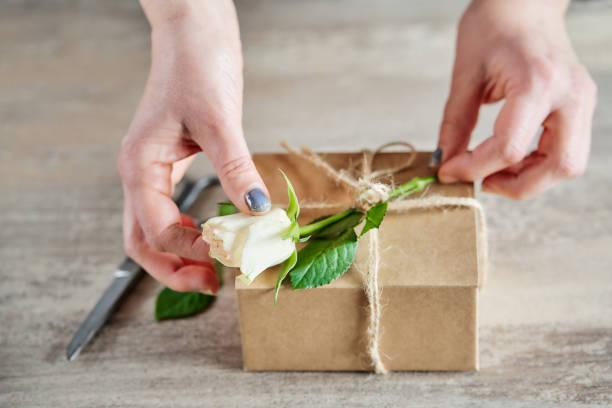 istock Female hands preparing a gift box with white rose on wooden background 1390324473