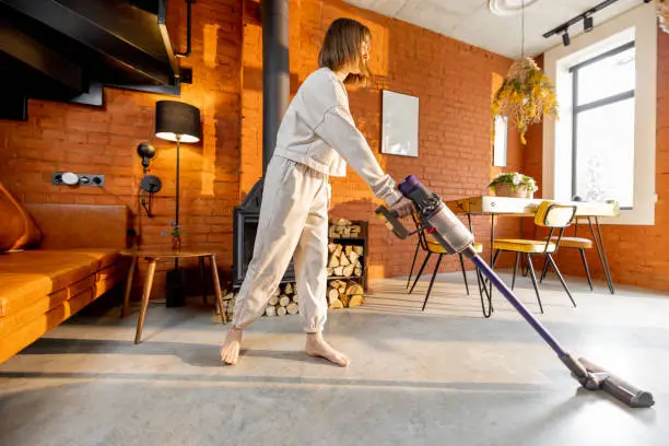 Woman cleaning concrete floor with cordless handheld vacuum cleaner in living room at home. Housewife doing housework