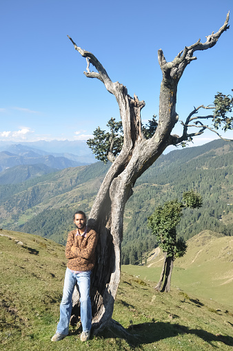 Full length of a good looking young guy looking at camera posing with crossed arms while leaning against tree in hilly region