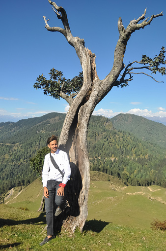 A young male tourist posing against tree with beautiful mountain and sky view