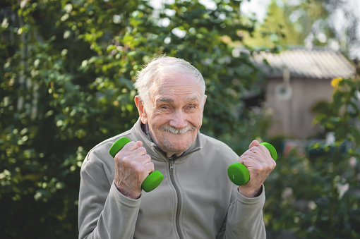 old man with dumbbells. The fitness of the person engaged in in the garden. Active old age, healthy lifestyle.