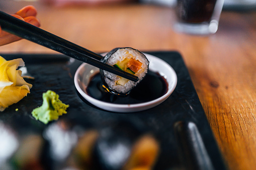 Hand holding chopsticks with sushi roll while soaking it in soy sauce