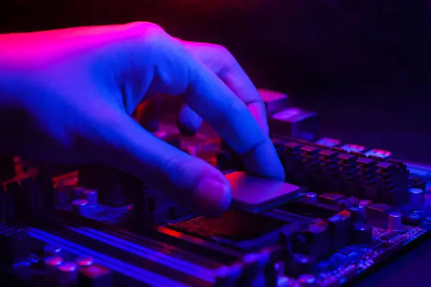 Photo of Motherboard in neon light. A hand inserts a processor into a socket on the motherboard. Computer service.