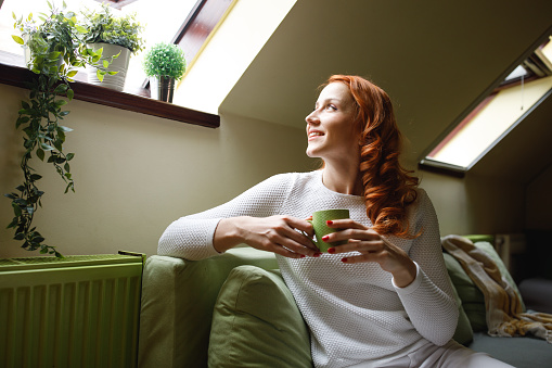 Copy space shot of happy young woman lounging on the sofa in her living room, looking through the window, daydreaming and enjoying a cup of coffee.