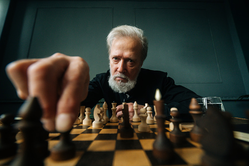 Close-up low-angle view of serious gray-haired mature male performing move with pawn piece on wooden chessboard, looking at camera. Handsome bearded man playing chess alone at home.