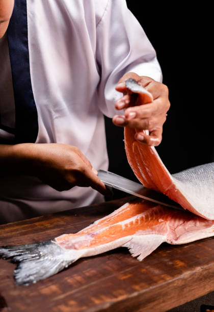 mano dello chef che tiene un pezzo di salmone fresco. primo piano delle mani dello chef che preparano cibo giapponese. chef giapponese che prepara sushi al ristorante. chef che prepara sushi tradizionale giapponese su tavola di legno. - thai cuisine asian cuisine vertical close up foto e immagini stock