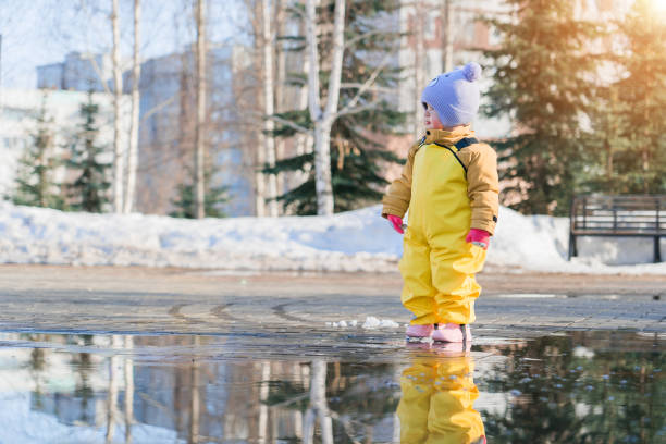 ребенок в возрасте 2-3 лет, одетый в желтый резиновый комбинезон, весной стоит в луже в городском парке - child looking messy urban scene стоковые фото и изображения