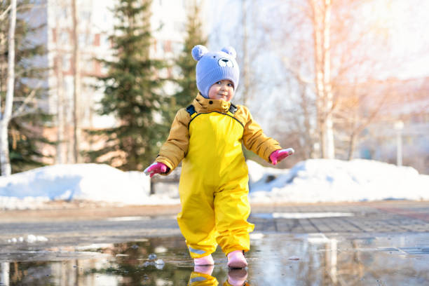 милый малыш 2-3 лет, одетый в желтый водонепроницаемый комбинезон, идет по луже в весеннем парке и улыбается - child looking messy urban scene стоковые фото и изображения