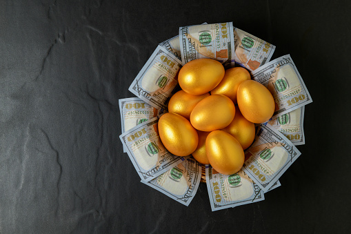 golden eggs and dollars in a basket on black background, top view