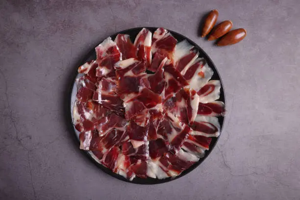 Ration of 100% Dehesa de Extremadura acorn-fed Iberian ham on a black plate, on a dark gray table and decorated with acorns