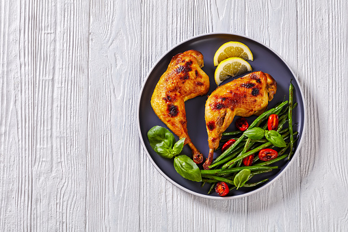 roast chicken legs with green beans, lemon, fried tomatoes and fresh basil on plate on white  textured wooden table, horizontal view from above, flat lay, copy space
