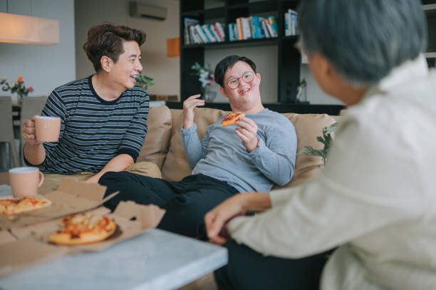 asiatische chinesische mehrgenerationenfamilie mit down-syndrom-enkel, der am wochenende pizza fast food im wohnzimmer auf dem sofa genießt - grandmother senior adult child multi generation family stock-fotos und bilder