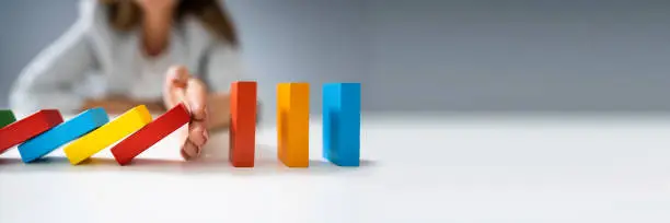 High Angle View Of A Businessperson Stopping Colorful Dominoes From Falling On Desk