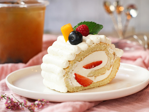 Close up of swiss roll cake filling with vanilla cream decorated with a pieces of assorted fresh fruits such as strawberry, mango, blackcurrant raspberry, in a white plate