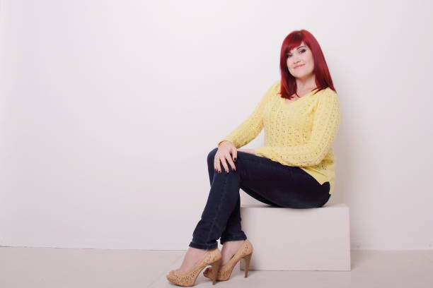 istock Red hair woman in jeans siting on white cube, studio shooting, copy space 1390479605