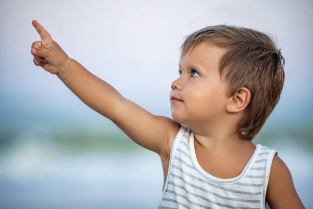 kind in einem gestreiften t-shirt mit blauen großen shows in die ferne vor dem hintergrund des meeres mit wellen - baby pointing child showing stock-fotos und bilder