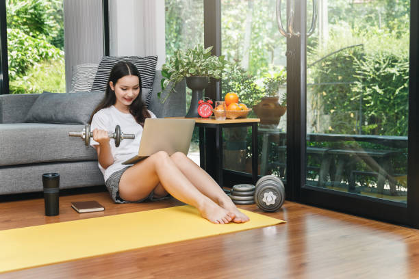 Beautiful young woman with dumbbell watching fitness video tutorials online on laptop while sit down on floor in living room. stock photo
