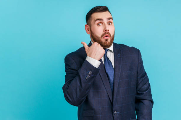 istock Astonished bearded man wearing dark suit pointing finger aside, showing copy space for commercial text, blank wall with idea presentation. 1390494270
