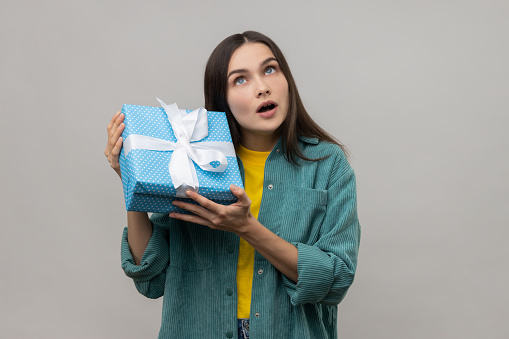 Portrait of curious woman shaking wrapped present on birthday, trying to guess what it is, looks doubtfully up, wearing casual style jacket. Indoor studio shot isolated on gray background.