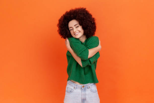 Woman with Afro hairstyle wearing green casual sweater hugging herself and smiling, feeling comfortable and fulfilled, narcissistic egoistic person. stock photo