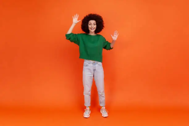 Full length portrait of woman with Afro hairstyle wearing green casual style sweater standing with raised arms, waving hello, glad to meet you. Indoor studio shot isolated on orange background.
