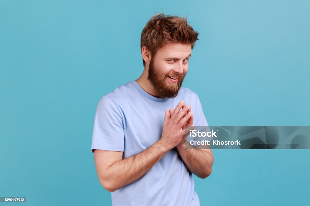 Man in T-shirt schemes something keeps hands together makes plans, sly expression looks thoughtfully Portrait of cunning bearded man schemes something keeps hands together and makes plans, sly expression looking at camera thoughtfully. Indoor studio shot isolated on blue background. Men Stock Photo
