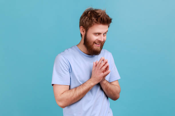 Man in T-shirt schemes something keeps hands together makes plans, sly expression looks thoughtfully Portrait of cunning bearded man schemes something keeps hands together and makes plans, sly expression looking at camera thoughtfully. Indoor studio shot isolated on blue background. smirking stock pictures, royalty-free photos & images