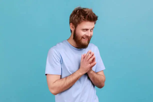 Photo of Man in T-shirt schemes something keeps hands together makes plans, sly expression looks thoughtfully