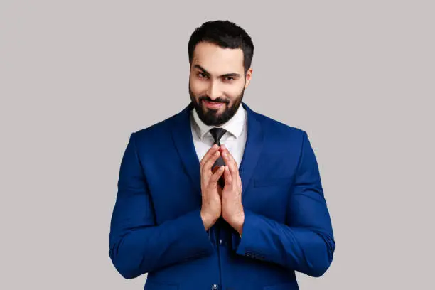 Cunning bearded man clasping hands and planning evil tricky prank or scheming, cheating with sly smile, wearing official style suit. Indoor studio shot isolated on gray background.