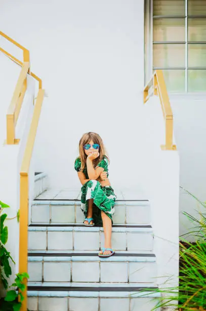 Photo of Sassy stylish little girl in a tropical outfit and sunglasses  sits waiting impatiently on the steps of a Miami Beach apartment building