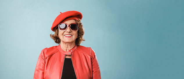 Senior woman wearing red leather jacket, sunglasses and beret smiling while posing over an blue background.