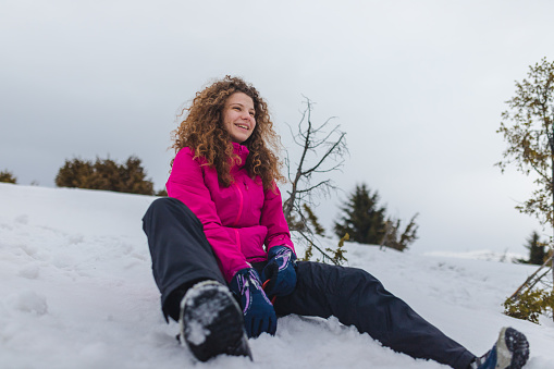 A beautiful young woman sliding down a snowy hill while smiling. She has curly brown hair.