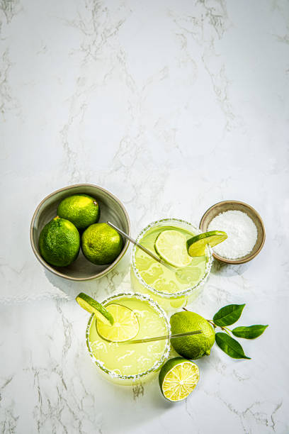 This is a photograph of a modern margarita glass with a rim of salt surrounded by fresh cut limes photographed on a white marble background