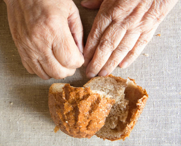 An old woman holds stale bread in her hands. Wrinkled hands of an old woman and a loaf of rye bread.A piece of dry bread on the table. The concept of social assistance to the poor. Hunger, poverty, loneliness.